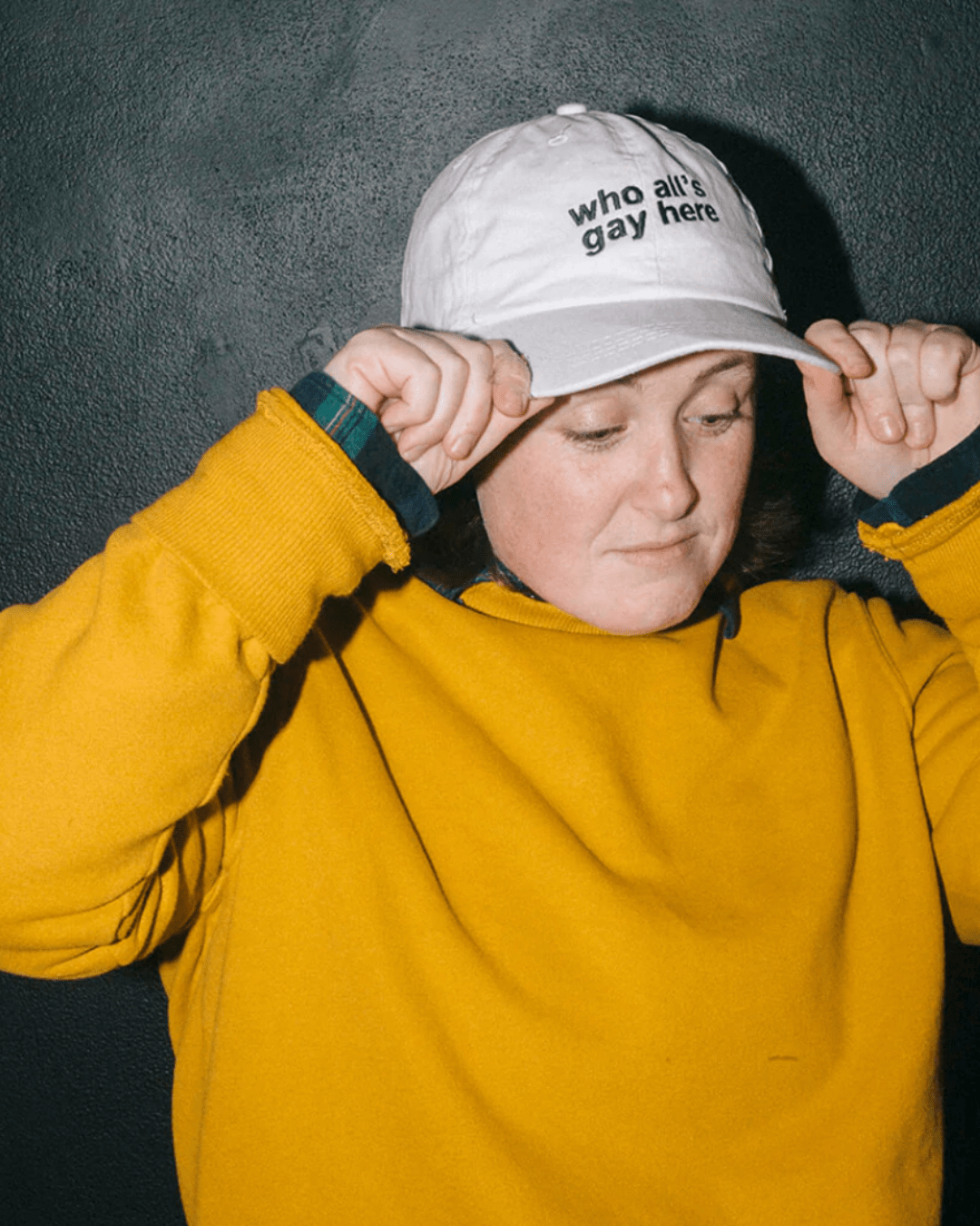 A model is wearing the Who All's Gay Here Hat. The hat is a baseball cap style in white color with black sans-serif text that says "Who All's Gay Here." The text is askew at a diagonal in two lines. The design is by Erin Sullivan.