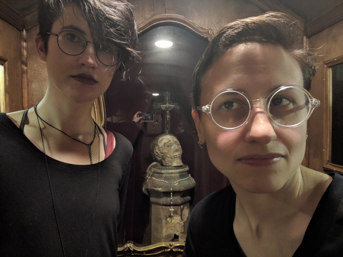 Nico, a white genderqueer human here with dark short hair that is shaved on the sides and glasses and a black shirt stands with sadie a white butch woman with glasses and short brown hair. they are standing in front of an art piece that looks like a reliquary with a skull in it.