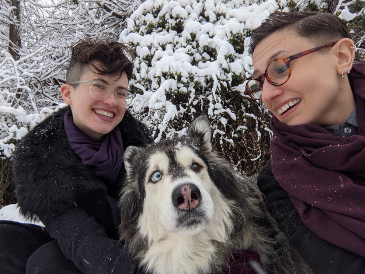  in the photo, sadie, nico and mya the dog pose in the snow. nico is a white genderqueer human with short dark hair that is shaved on the sides and glasses. they are wearing a black coat. sadie is a white butch woman with short hair and glasses who is also wearing a black coat and a burgundy scarf. mya is a fluffy white, gray and brown malamute mix with two different colored eyes