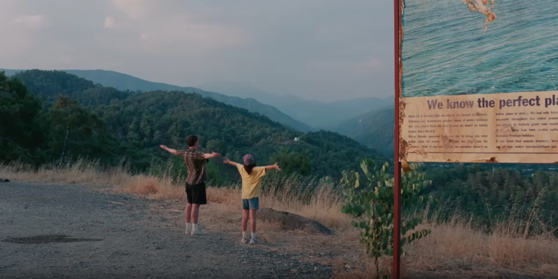 A father and daughter stand on a cliff in Turkey with their arms outstretched. A nearby sign reads "We know the perfect place"