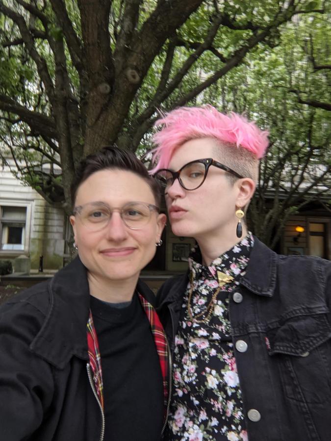 nico kisses sadie on the cheek. sadie is wearing a Harrington jacket and Nico is wearing a floral print shirt (the same one sadie was wearing in the previous picture) and also has their hair dyed hot pink