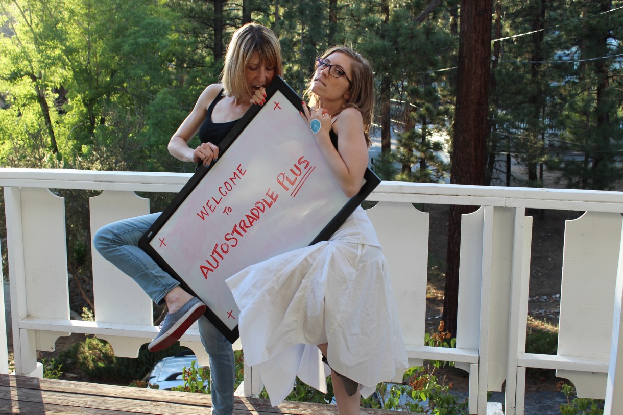 Laneia and Riese holding a whiteboard sign that says "Welcome to Autostraddle Plus"
