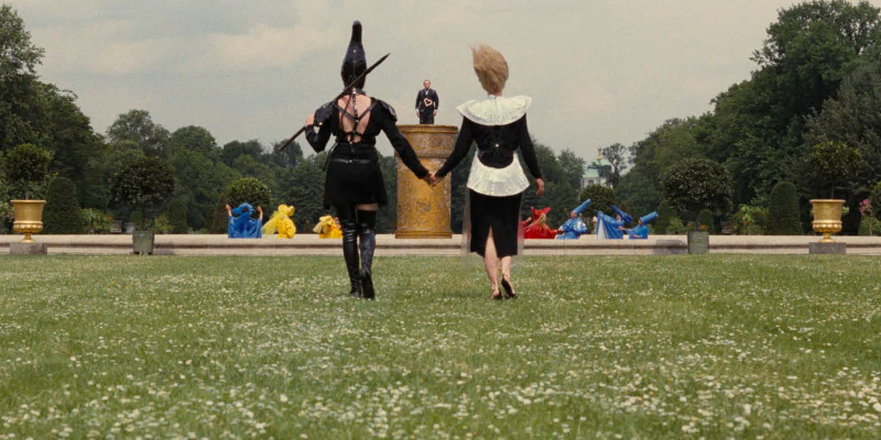 Two women wearing outlandish outfits hold hands in a grassy field and look up at a man standing on a pillar. 