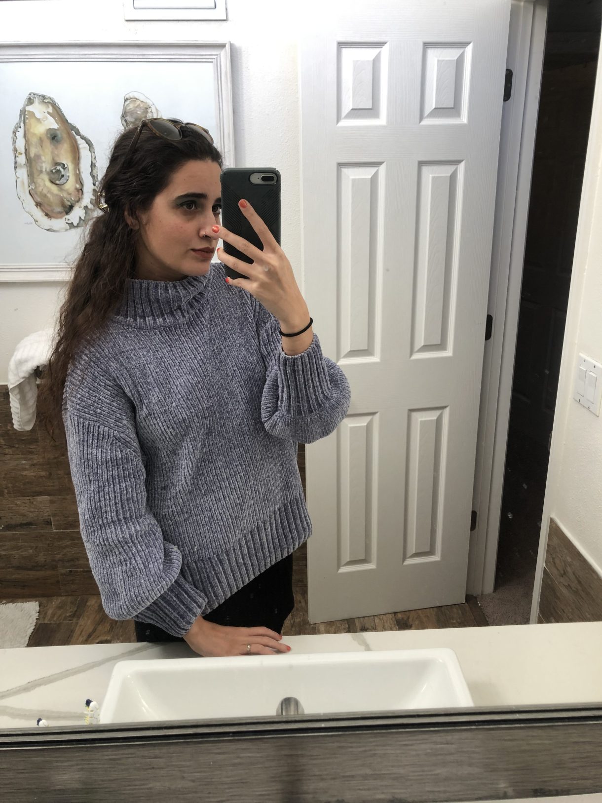Kayla Kumari Upadhyaya wears a chunky blue-gray turtleneck sweater over a black skirt. Her hair is half up and she has sunglasses on her head.