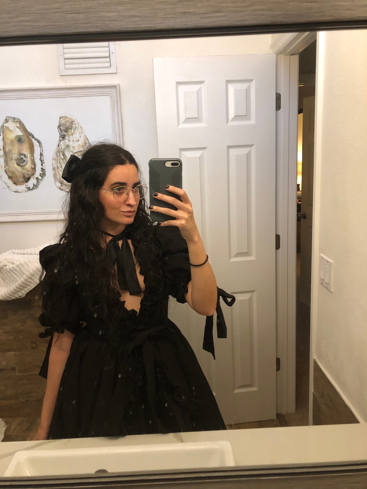 Kayla Kumari Upadhyaya wears a low V-neck black frilly haunted dollesque dress with a ribbon on her neck and in her hair which is parted in the middle. She wears gold framed glasses and her nails are painted black.