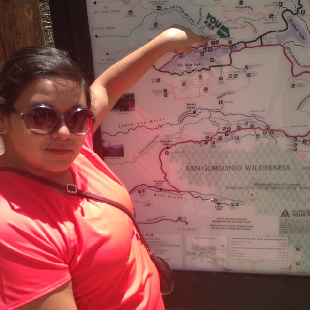Yvonne in a orange t-shirt and sunglasses, pointing at a trail map