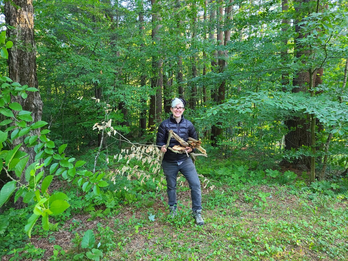 Nico stands in a jacket and jeans and hiking boots at the edge of a forest, holding a bundle of sticks and smiling.