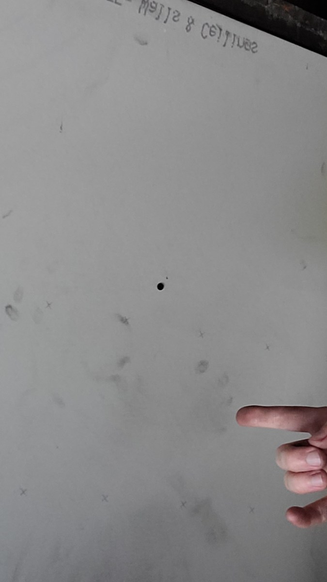 a photo that is mostly of some newly installed drywall on a ceiling with nico, a white genderqueer human partially visible at the bottom, lifting up one dirty hand and giving the horns with it and peering at the camera through glasses. they are wearing an orange trucker hat.
