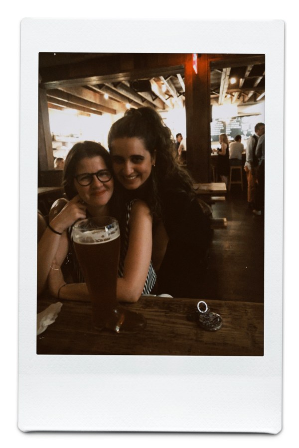 Kayla hugs a sitting Kristen from behind in a German bar, and there's a boot full of beer on the table