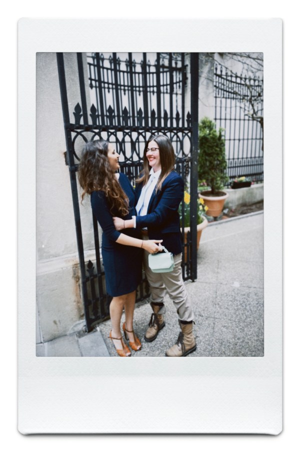 Kristen in a blue suit and khakis holds Kayla in a blue dress in front of an ornate gate, and they're looking at each other and laughing