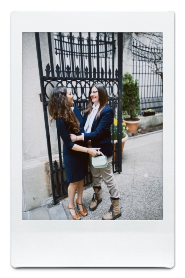 Kristen in a blue suit and khakis holds Kayla in a blue dress in front of an ornate gate, and they're looking at each other and laughing
