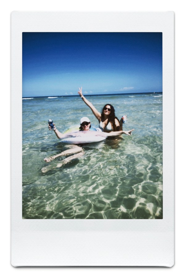 Kristen is in a floating intertube holding a drink and Kayla crouches in the ocean behind her holding a drink and throwing up a peace sign