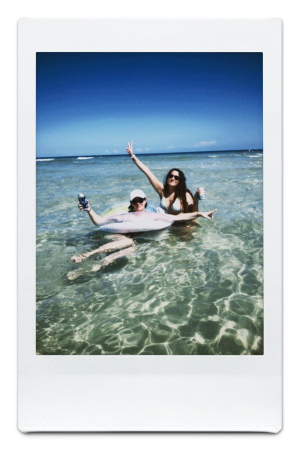 Kristen is in a floating intertube holding a drink and Kayla crouches in the ocean behind her holding a drink and throwing up a peace sign