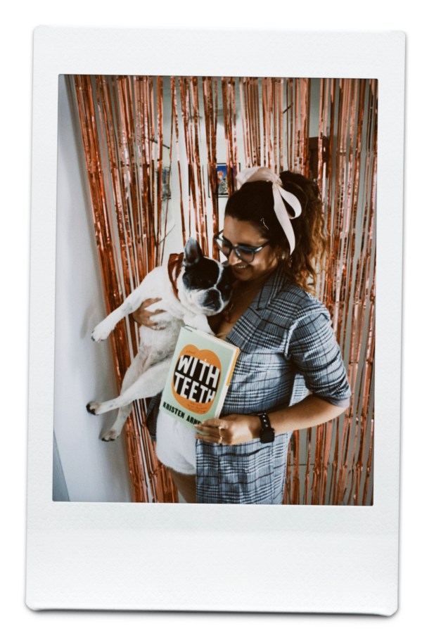 Kayla holds Kristen's book With Teeth as well as Lola the French bulldog and stands in front of pink streamers
