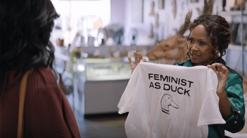 Crystal Fox as Izzy in The Big Door Prize. She's holding up a shirt that says "Feminist as Duck."