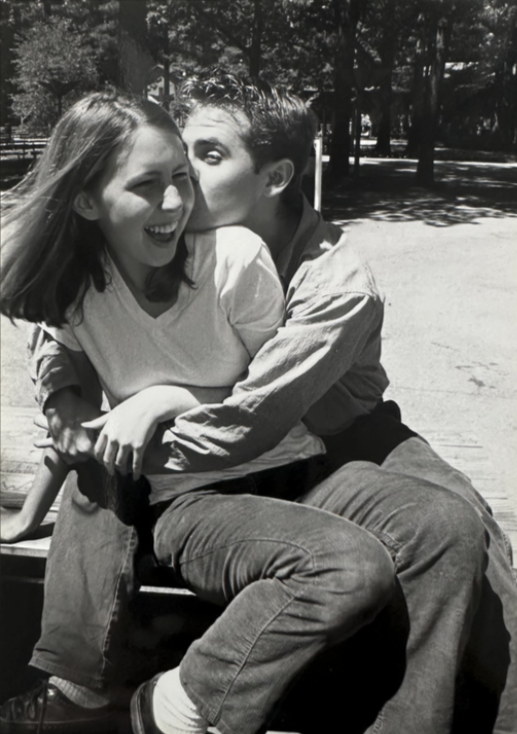 a black and white photo of Riese laughing while sitting wrapped up in the arms o fher gay boyfriend. they are outside.