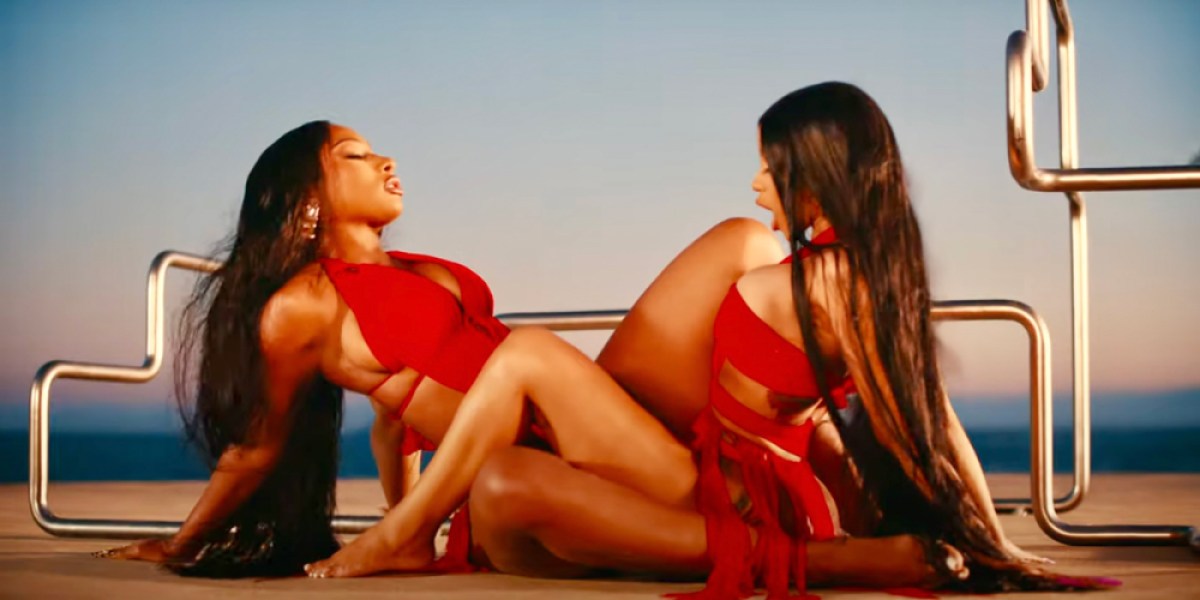 Cardi B and Megan Thee Stallion are in red bathing suits with their legs interlocked together on a pool deck.