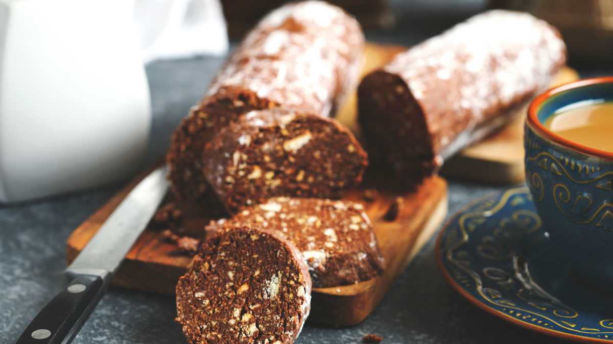 A partly cut up chocolate salami on a cutting board next to a cup of coffee.